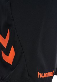 Short de sport noir avec deux chevrons orange sur le côté gauche et le mot "hummel" en orange près de l'ourlet inférieur.