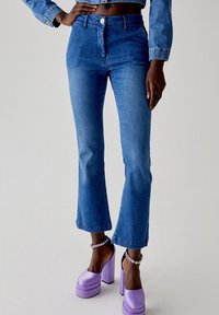 Jeans flare in denim blu con vita alta, dotati di chiusura con bottone e zip, e orli rifiniti, abbinati a scarpe con piattaforma viola.