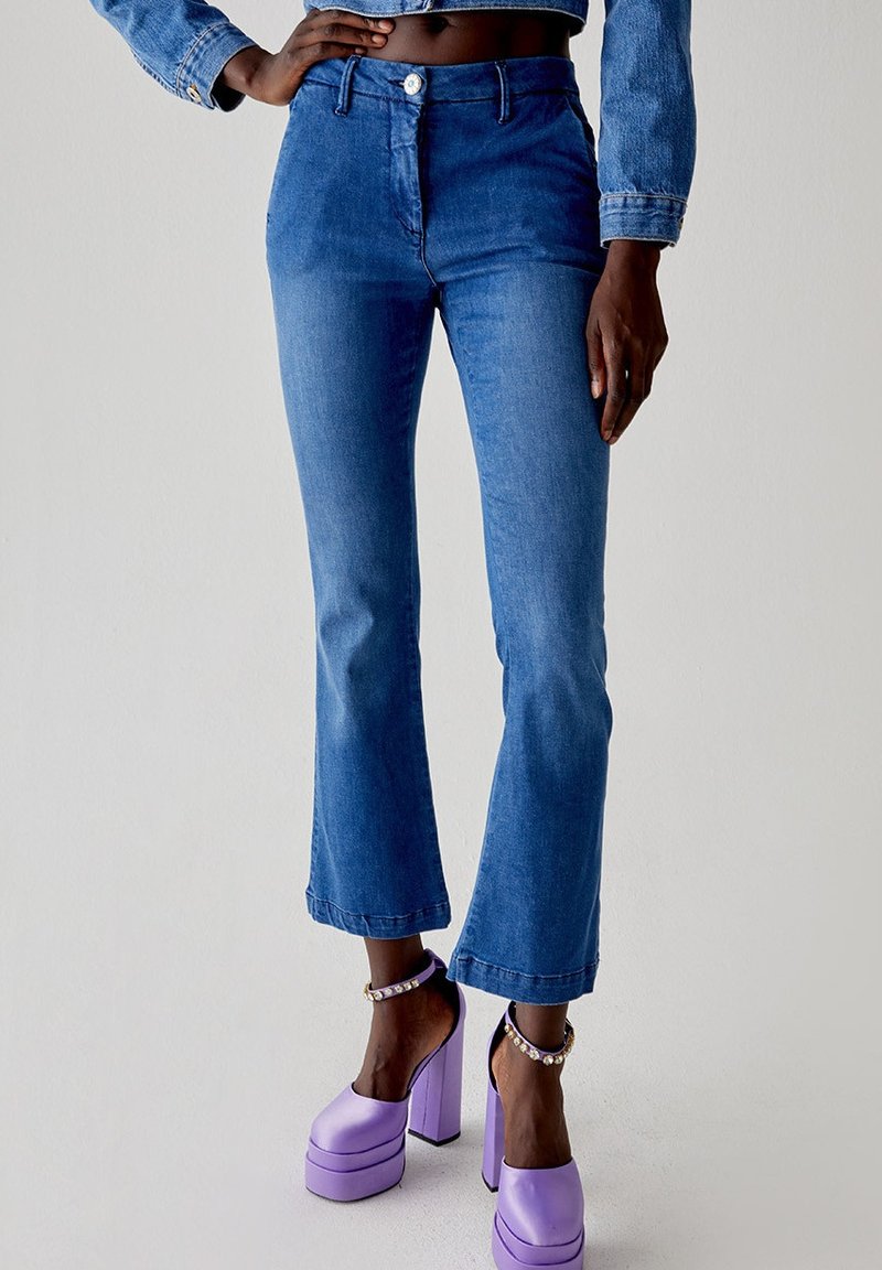 Jeans flare in denim blu con vita alta, dotati di chiusura con bottone e zip, e orli rifiniti, abbinati a scarpe con piattaforma viola.