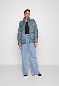 Casaco acolchoado azul-petróleo com gola alta, fecho de correr frontal e detalhes em preto, combinado com jeans de pernas largas azul claro e ténis pretos.