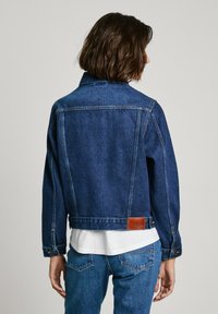 Giacca di jeans blu scuro dal design classico, con una toppa in pelle sul retro, polsini con bottone e lunghezza corta.