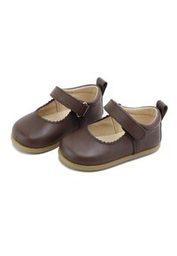 Zapatos Mary Jane de cuero marrón con bordes ondulados, una correa y hebilla, suelas planas de goma y un forro interior liso.