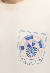 Αφηρημένος κυκλικός μπλε και μπεζ σχεδιασμός με σχήμα λουλουδιού και το κείμενο "Scotch & Soda" σε ανοιχτό μπεζ ύφασμα.