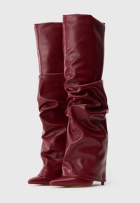 Botas de cuero burdeos altas y holgadas con punteras puntiagudas y tacones bajos y finos contra un fondo blanco liso.