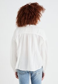 White, loose-fitting blouse with textured fabric, gathered details at shoulders, and elastic cuffs, paired with light blue jeans.