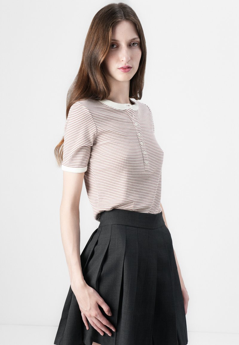 Polo w paski z miękkiej tkaniny z zapięciem na guziki, w połączeniu z plisowaną czarną spódnicą. Koszulka ma kontrastujący biały kołnierzyk.