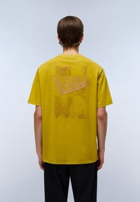 Gul bomulds T-shirt med en stor, tekstureret rød "NAPAPIJRI" grafik på ryggen, korte ærmer og rund hals.