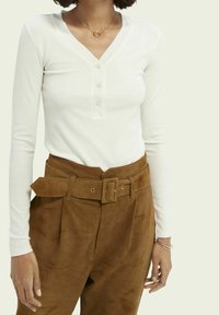 Witte geribbelde lange mouwen top met een v-hals en vier knopen, gecombineerd met hoge taille bruine suède broek met een riem.