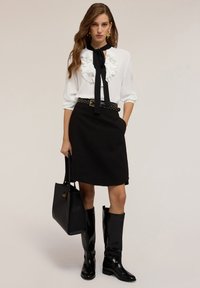 Blusa bianca con parte anteriore arricciata e cravattino nero, abbinata a una gonna nera, cintura nera con borchie, stivali neri fino al ginocchio e borsa.