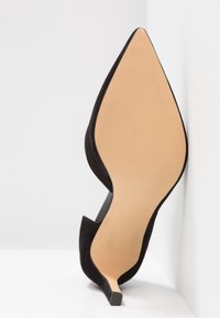 Chaussure à talon haut noire avec bout pointu et semelle beige, positionnée droite contre un mur d’angle blanc et le sol.