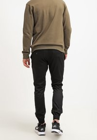 Pantalons de jogging noirs avec des poignets élastiques, coupe slim et poches latérales, associés à un sweat à capuche vert et des baskets noires. Texture lisse.
