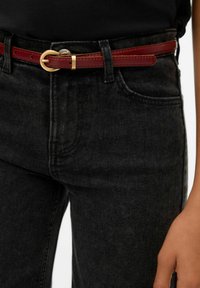 Vinrött skärp i läder med ett guldfärgat cirkulärt spänne, bärs över svarta jeans och har en jämn struktur samt synliga sömmer.