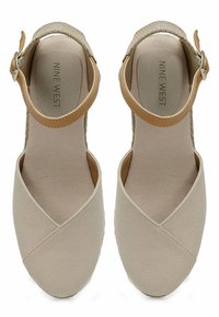 Beige canvas sandalen met een kruispatroon, verstelbare enkelband en een gestructureerde binnenzool. Merkennaam "NINE WEST" is ingedrukt aan de binnenkant.