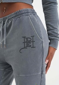 Grå sweatpants av mjukt tyg, med elastisk midja, sidoficka och svart grafisk logotyp på framsidan.