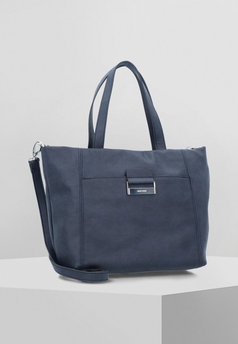 Sac cabas en tissu bleu marine avec deux poignées courtes, une bandoulière longue amovible et une plaque métallique à l'avant, posé sur un socle blanc.
