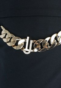 Cintura in catena d'oro con grandi link intercambiabili e una chiusura decorativa con una 'J' adornata di strass. Sfondo in tessuto scuro.