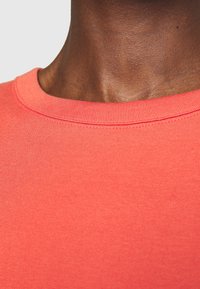Coral-colored crew neck t-shirt with ribbed neckline. Smooth cotton texture, featuring subtle stitching details. Close-up view from neck area.