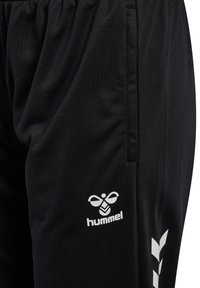 Gros plan sur un pantalon de sport noir avec une ceinture élastique, une poche latérale, un logo Hummel blanc et un motif chevron sur la jambe droite.