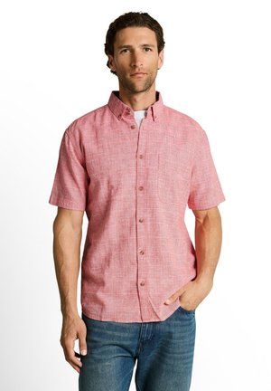 Mann trägt ein kurzärmeliges, pinkfarbenes Hemd mit Brusttasche und blaue Jeans, steht mit einer Hand in der Tasche.
