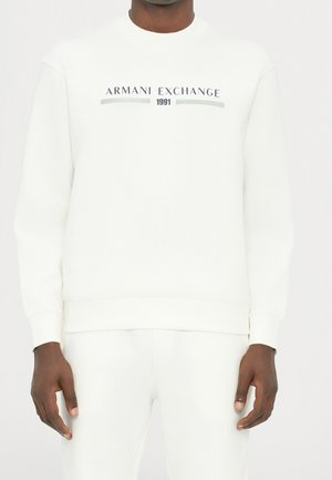 Homme portant un sweat-shirt blanc Armani Exchange avec le texte « 1991 » et un pantalon blanc assorti, visible du cou aux hanches.