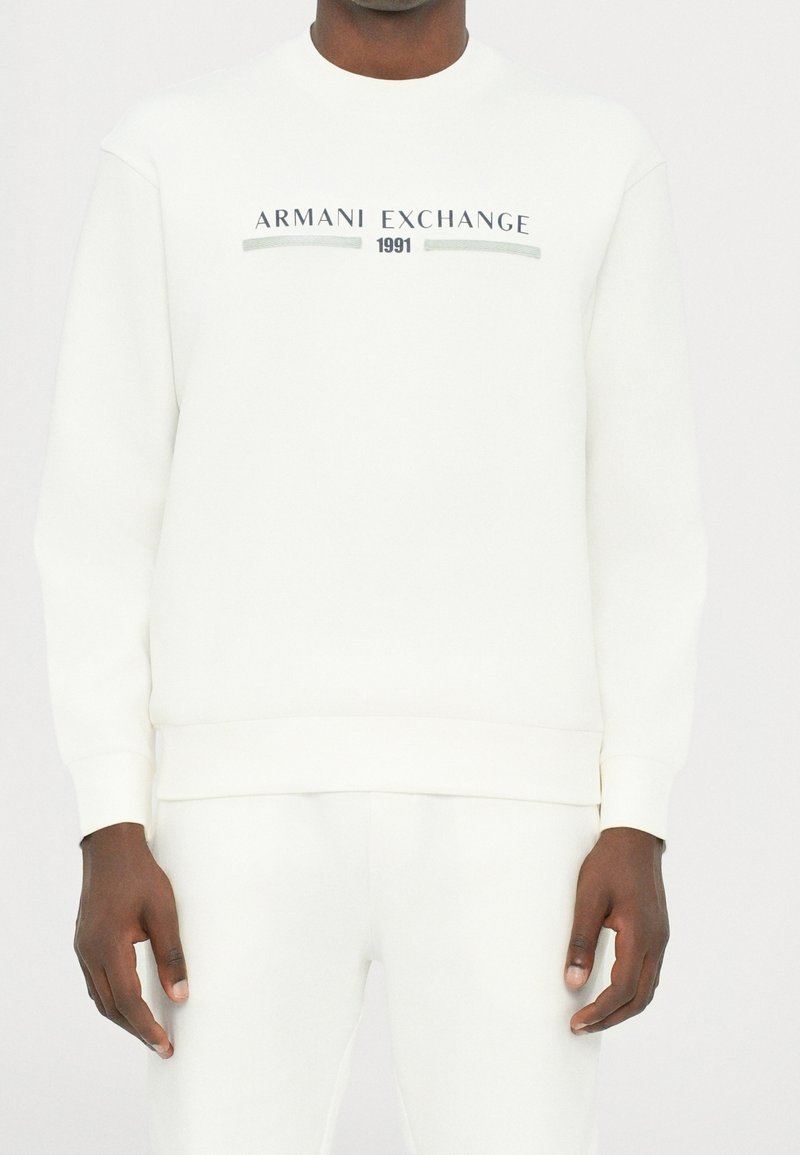 Homme portant un sweat-shirt blanc Armani Exchange avec le texte « 1991 » et un pantalon blanc assorti, visible du cou aux hanches.