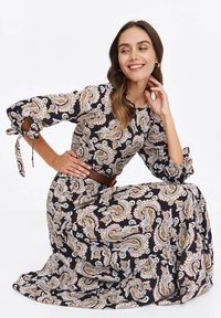 Woman kneeling, smiling, wearing long paisley dress with tied sleeves and brown belt, against white background.