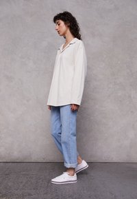 Camicia in cotone crema oversize con colletto, abbinata a jeans in denim blu chiaro arrotolati ai polsini e sneakers bianche con dettagli rossi.