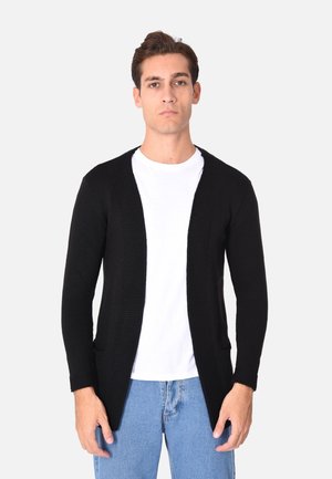 Cardigan nero a maglia con apertura frontale, maniche lunghe e due tasche anteriori, abbinato a una t-shirt bianca e jeans blu.