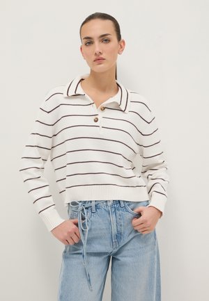 Jonge vrouw met donker haar in een staart draagt een wit polotrui met lange mouwen met dunne donkere strepen en een lichtblauwe jeans met hoge taille.