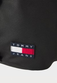 Sort tekstil med en struktureret overflade, udstyret med en broderet mærkat med "TOMMY JEANS" og et rødt, hvidt og blåt logo.