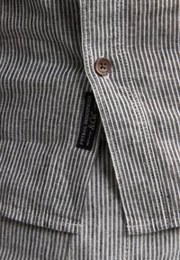 Chemise à boutons rayée présentant un mélange de lignes gris foncé et blanches, avec un bouton brun et une étiquette indiquant "Petrol Industries".