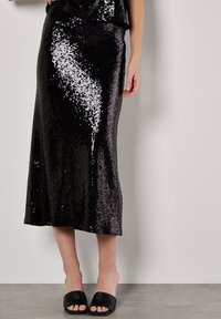 Zwarte pailletten midi-rok met een semi-aangesloten ontwerp, met een soepele textuur en subtiele glans. Gepaard met open teen zwarte instapsandalen.