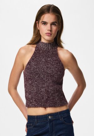 Jeune femme portant un crop top en maille bordeaux sans manches à col montant et un jean bleu foncé, debout les mains dans le dos.