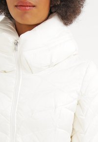 Vit kviltad pufferjacka med hög krage och fullt blixtlås, med en slät yta och subtila diagonala sömmar.