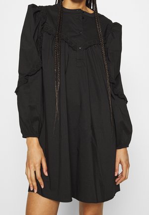 Woman wearing a loose black dress with long puffed sleeves, buttoned yoke, and lace trim on shoulders and sleeves.