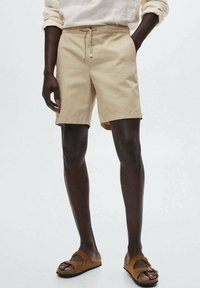 Pantaloni corti in cotone beige con vita elastica e cordino. Abbinati a sandali marroni da infilare e una camicia leggera e dalla vestibilità ampia.