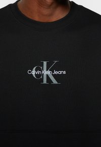 Svart sweatshirt med rund halsringning och ribbade muddar, med en kontrasterande broderad "CK"-logotyp och texten "Calvin Klein Jeans" på bröstet.
