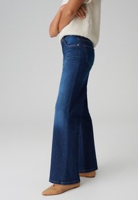 Dunkelblaue Jeans mit weit ausgestelltem Bein aus Denim, mit mittelhoher Taille, Seitentaschen und einem sauberen Saum. Kombiniert mit nudefarbenen Ballerinas.