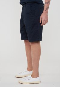 Pantaloni corti in cotone blu navy con tasche laterali, chiusura a bottone e orli arrotolati. Indossati con sneakers bianche in tela.