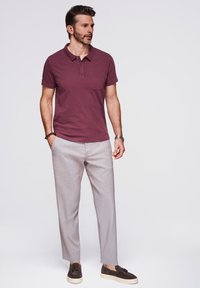 Polo a manches courtes color bordeaux con taschino, abbinato a pantaloni grigi chiari. Scarpe slip-on marroni con finitura texturizzata.