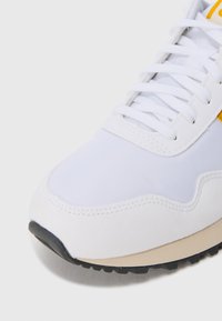 White sneaker front with white laces, beige midsole, black outsole, and a small yellow detail visible on the side.
