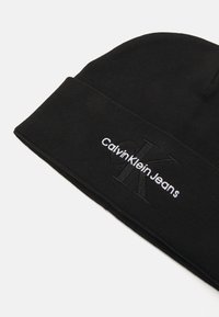 Fekete kötött sapka behajtott széllel, amelyen fehér "Calvin Klein Jeans" logó és finom kiemelt betűs részletezés található az elején.