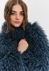 Na dotyk mäkký, modro-čierny kožuch s huňatou textúrou, dlhé rukávy a vysoký golier, voľne obopínajúci telo nositeľa.