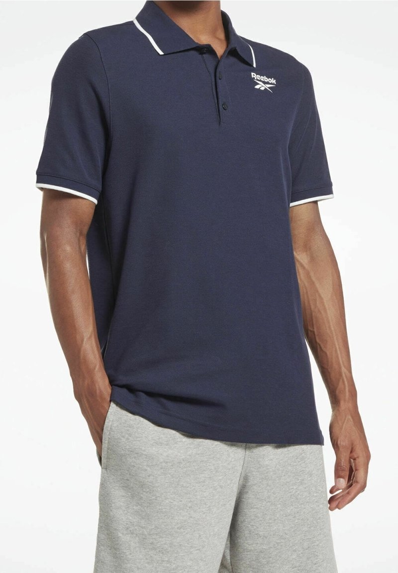 Námořnická modrá polo košile s bílým límcem a lemy na rukávech, s logem Reebok na hrudi. Vyrobena z hladkého, prodyšného materiálu.