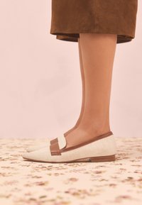 Mocassins en tissu beige avec des accents en cuir marron, un bout pointu, un petit talon en bois et un détail de bride en cuir sur le dessus.