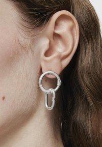 Pendientes de aro plateados que presentan formas circulares y ovaladas entrelazadas, con textura suave y un diseño minimalista.