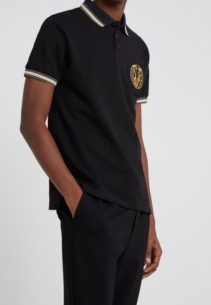 Polo noir avec emblème doré, col côtelé et accents rayés sur les manches. Confectionné dans un tissu doux, avec une coupe classique et des boutons.