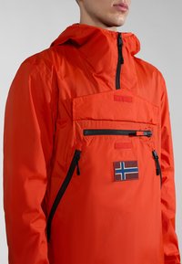 Heldere oranje waterdichte jas van nylon, voorzien van een voorzak met rits, halve rits sluiting en een patch met een vlag embleem.