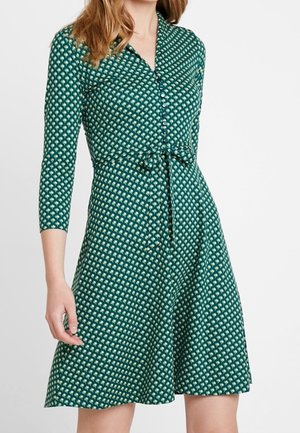 Femme portant une robe verte à motifs avec des manches trois-quarts, des boutons devant et une ceinture nouée à la taille.