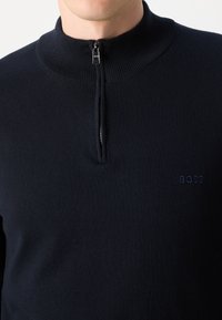Marinblå zip-up-tröja med hög krage och ribbad detalj. Har en diskret präglad logotyp på bröstet och är tillverkad av mjukt stickat tyg.